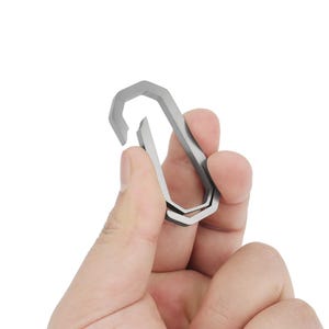 Può includere: Un moschettone in metallo argentato con un design unico e multiforme. Il moschettone è aperto e pronto per essere utilizzato.