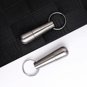 May include: Two stainless steel capsule-shaped keychains with silver key rings. The capsules have a rounded end and a screw-on top. One keychain is on a black textured surface, and the other is on a white surface.