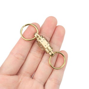 Peut inclure: Un porte-clés de couleur laiton avec deux anneaux et un composant central cylindrique. Le composant comporte une série de petits trous espacés. Les anneaux et le composant central sont reliés. Le porte-clés est tenu dans une main.
