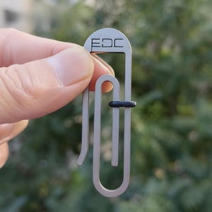 May include: A silver-coloured EDC paperclip-style tool with the letters "EDC" etched on the top. The tool has a curved design with a black rubber ring. The background is a blurred green.