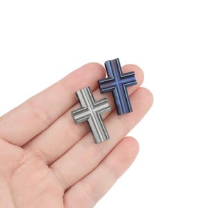 May include: Two metal cross pendants, one silver and one blue, are held in a hand. The crosses have a detailed design and are perfect for jewelry making.