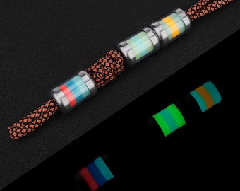 May include: A close-up of a lanyard featuring three cylindrical beads with a stainless steel finish and colourful stripes. The lanyard is made of a brown and black braided cord. The beads glow in the dark, showing green, blue, and orange.