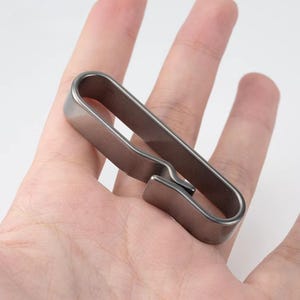 May include: A silver-coloured metal clip, possibly for holding money or papers. The clip has a simple, rectangular design with a curved top and a small, angled opening. The object is held in a person's hand.