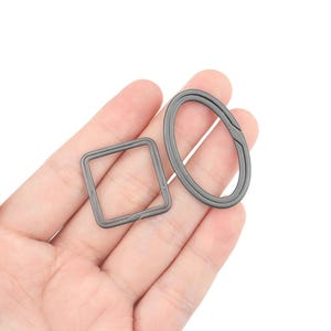 Puede incluir: Dos llaveros de metal, uno ovalado y otro cuadrado, en una mano. Los anillos son ambos de color plateado.