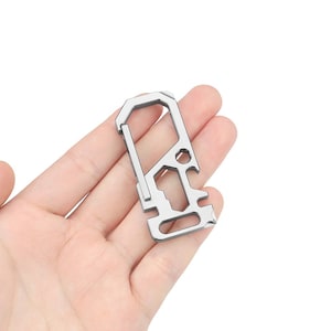 Puede incluir: Un mosquetón multiherramienta plateado con varias herramientas integradas. El mosquetón tiene forma rectangular con una puerta con bisagras y varias secciones recortadas. Parece estar hecho de metal y está diseñado para el uso diario.
