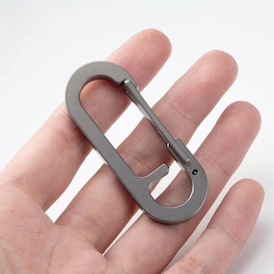 Op de afbeelding: Een grijze metalen karabijnhaak met een gebogen ontwerp. De haak wordt in een hand gehouden en heeft een veergeladen sluiting voor een veilige sluiting. De karabijnhaak is een veelzijdig hulpmiddel.