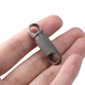 Puede incluir: Pequeño accesorio de llavero gris metálico con un agarre texturizado con patrón de diamantes. El accesorio tiene dos aberturas circulares en cada extremo. Se sostiene en una mano sobre un fondo blanco. El artículo parece ser de metal.
