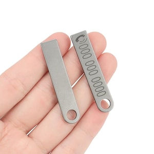 Könnte beinhalten: Zwei silberfarbene rechteckige Anhänger mit abgerundeten Kanten. Einer hat ein Telefonsymbol und eine eingravierte Telefonnummer. Der andere ist leer. Beide haben ein kleines Loch an einem Ende.