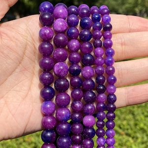 May include: A strand of purple amethyst beads with a variety of shades and patterns. The beads are round and smooth, and they are strung on a clear elastic cord.