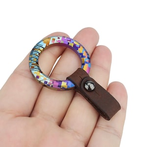 Può includere: Un portachiavi in titanio iridescente multicolore con un cinturino in pelle marrone e un bottone nero.