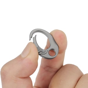 Könnte beinhalten: Ein kleiner, silberfarbener Karabinerhaken mit gebogenem Design. Der Haken hat einen federbelasteten Verschluss und ein dekoratives Muster in der Nähe der Öffnung. Der Karabiner wird zwischen zwei Fingern vor weißem Hintergrund gehalten.