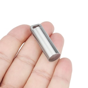 Pode incluir: Um pequeno recipiente cilíndrico de metal prateado, com uma base arredondada e uma alça retangular na parte superior. O recipiente é segurado na mão de uma pessoa e parece ser feito de aço inoxidável.