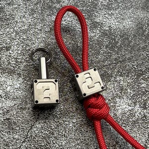 May include: A silver key chain with a question mark block design. The key chain is attached to a red rope.