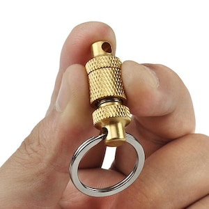 May include: A brass-colored keychain accessory with a textured, cylindrical body and a silver-colored key ring. The accessory has a swivel design and is held in a person's hand against a white background.