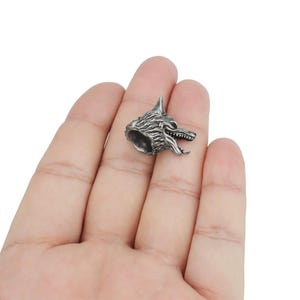 Puede incluir: Un pequeño colgante plateado con forma de cabeza de lobo con detalles de pelaje y boca abierta, mostrando dientes. El colgante es probablemente de metal y está diseñado para ser usado como joya o en manualidades.