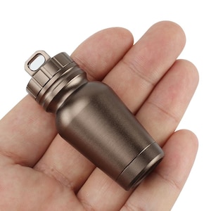 Puede incluir: Una pequeña botella marrón metálica con tapa de rosca y un lazo para sujetarla a un llavero. La botella tiene un cuerpo cilíndrico y una base redondeada. El diseño es minimalista y funcional.