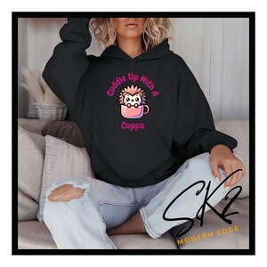 Op de afbeelding: Zwarte hoodie met een roze afbeelding van een egel in een roze mok. De afbeelding zegt "Cuddle Up With A Cuppa".