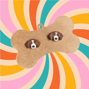 Pode incluir: Um par de brincos com a forma de rosto de cachorro marrom e branco em um cartão marrom em forma de osso contra um fundo colorido de listras laranja, amarelas, rosa e azuis que giram.