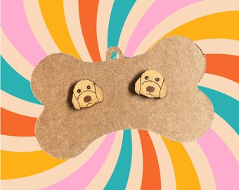Può includere: Un paio di orecchini in legno con un design a forma di viso di cane carino. Gli orecchini sono di colore marrone chiaro con uno sfondo rosa chiaro e sono esposti su un pezzo di cartone a forma di osso.