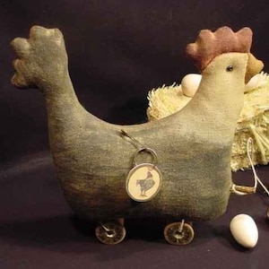 Könnte beinhalten: Ein graues und braunes Stoff-Hühner-Spielzeug mit Rädern, das einen kleinen Wagen mit Heu zieht. Das Huhn hat einen roten Kamm und einen braunen Schnabel. Der Wagen ist mit einer kleinen braunen Spule und weißen Eiern verziert.