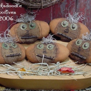 May include: Five handmade burlap pumpkins with painted faces and twig noses in a wooden bowl. The pumpkins are decorated with green buttons for eyes and have a rustic, primitive style. The bowl is filled with straw and there is a red leaf in the bowl.