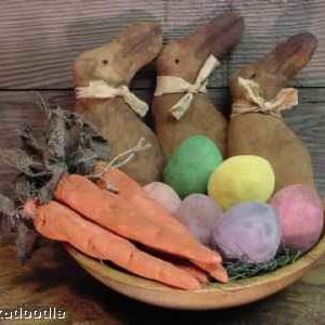 以下が含まれることがあります： カラフルなイースターエッグ、ニンジン、そして素朴な木製のウサギの置物3体が、木製のボウルに入っています。ウサギは茶色で、首には布のリボンが結ばれています。