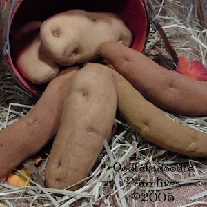 Könnte beinhalten: Handgefertigte Stoffkartoffeln in verschiedenen Braun- und Beigetönen, einige fallen aus einem rot umrandeten Korb. Die Kartoffeln sind auf Stroh angeordnet, was einen rustikalen, ländlichen Stil ergibt. Ein charmantes, handgefertigtes Produkt.