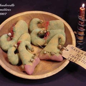 May include: A wooden bowl holds several green and pink elf shoe ornaments with bells and red ribbon. A lit candle sits to the right. A tag reads "Have a Holly Jolly Christmas 1862". The bowl is labeled "Oodlekadoodle Primitives 2007".
