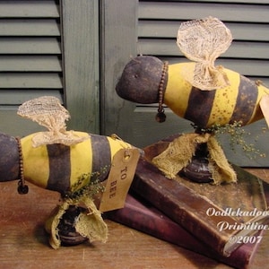 May include: Two handmade fabric bee figurines with black and yellow striped bodies, burlap wings, and a brown base. The bees are sitting on a stack of books with the text "Oodlekadoodle Primitives ©2007". One bee has a tag that says "To Bee" and the other has a tag that says "Not To Bee".