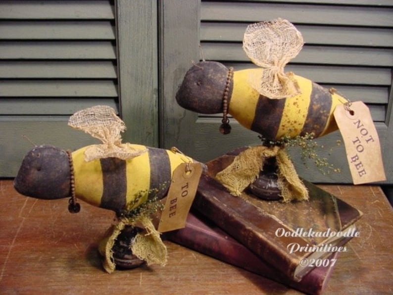 K&ouml;nnte beinhalten: Zwei handgefertigte Stoffbienchen mit schwarz-gelb gestreiften K&ouml;rpern, Fl&uuml;geln aus Sackleinen und einem braunen Sockel. Die Bienen sitzen auf einem Stapel B&uuml;cher mit dem Text "Oodlekadoodle Primitives &copy;2007". Eine Biene hat ein Schild mit der Aufschrift "To Bee" und die andere ein Schild mit der Aufschrift "Not To Bee".