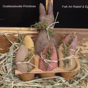 May include: A brown folk art rabbit with long ears sits in a cardboard egg carton with five painted eggs. The eggs are tied with twine and have sprigs of greenery on top. The carton is sitting on a bed of straw.