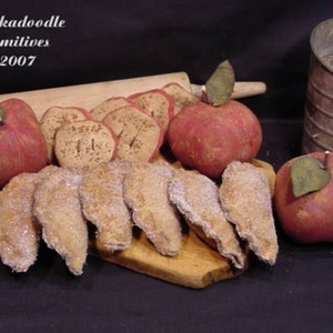 May include: A wooden cutting board with a variety of homemade apple pies, red apples, and a rolling pin. The text "Oodleka doodle Primitives ©2007" is visible in the top left corner of the image.