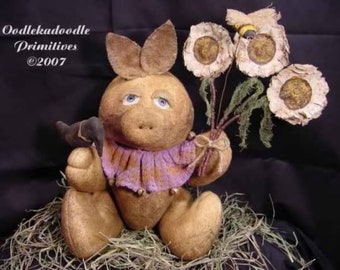 May include: A brown bunny doll with a purple sweater and blue eyes holds a small black bird in one hand and a bouquet of dried flowers in the other. The flowers are white with yellow centers and have a rustic, handmade look. The doll is sitting on a bed of straw. The text "Oodleka doodle Primitives ©2007" is visible in the top left corner of the image.