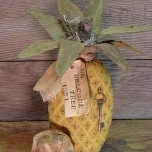 May include: A yellow pineapple-shaped decorative object with a brown burlap tag that says "We Welcome Thee 1st 11". The pineapple is sitting on a wooden surface.