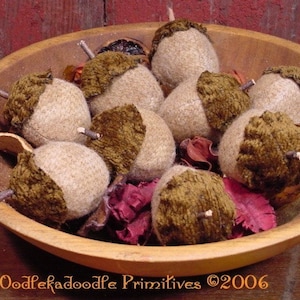 May include: A wooden bowl filled with handmade acorn decorations. The acorns have tan bodies and brown caps, with small twig stems. The bowl also contains dried red and brown decorative elements. The text "Oodlekadoodle Primitives ©2006" is visible.
