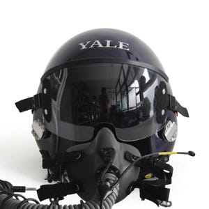 Könnte beinhalten: Ein marineblauer Pilotenhelm mit dem Wort "YALE" auf der Oberseite. Der Helm hat ein dunkles Visier, eine graue Sauerstoffmaske und verschiedene Riemen und Befestigungen. Der Helm ist für die Luftfahrt konzipiert.
