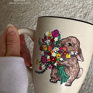 Puede incluir: Taza de cerámica beige con un conejo marrón que sostiene un ramo de flores coloridas. La taza tiene un borde negro y un asa blanca. El texto en la taza dice "finished work - selfmade for my mom".