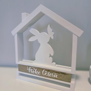 Könnte beinhalten: Weiße, holzerne Osterdekoration in Form eines Hauses mit einem Ausschnitt eines Hasen, der eine Blume hält. Die Basis des Hauses hat ein Holzschild mit der Aufschrift "Frohe Ostern" in weißen Buchstaben.