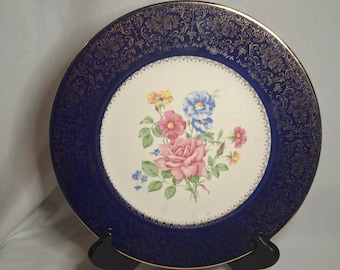 May include: A vintage blue and gold ceramic dinner plate with a floral design featuring pink, blue, and yellow flowers. The plate has a gold rim and a gold floral pattern around the edge.