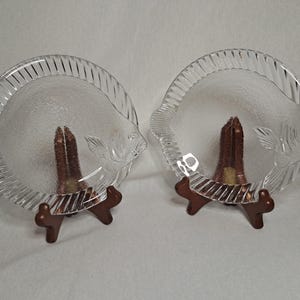 Könnte beinhalten: Zwei klare, fischförmige Servierplatten aus Glas mit strukturierter Oberfläche. Die Platten stehen auf Holzständern.