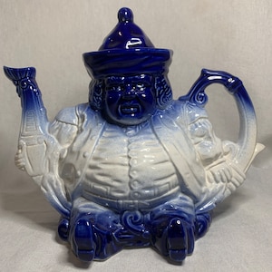 May include: A blue and white ceramic teapot shaped like a seated figure wearing a blue hat. The teapot has a spout shaped like a hand holding a scroll and a handle shaped like a hand holding a fan.