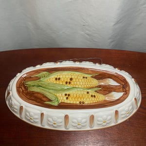 May include: Oval ceramic serving dish featuring two ears of corn. The corn is yellow with brown kernels and green husks, set against a brown background. The dish has a white border with floral and brown accents, perfect for kitchen decor.