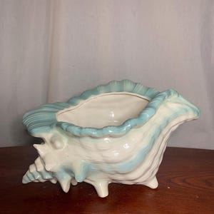 Peut inclure: Un pot de fleurs en céramique en forme de coquillage, blanc et bleu clair. Le pot a une grande ouverture et est soutenu par trois petits pieds. L'extérieur présente un motif texturé et strié avec une couleur d'accent bleu clair.
