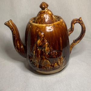 May include: A brown glazed ceramic teapot with a lid and a handle. The teapot has a raised design of a person and a dog.
