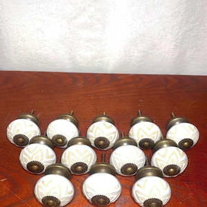 May include: Twelve white ceramic cabinet knobs with a cream floral pattern. Each knob features a bronze base and screw. The knobs are displayed on a dark wooden surface. Each knob measures approximately 4 cm in diameter.