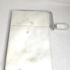May include: White marble cheese board with a chrome wire cutter and a white plastic handle.