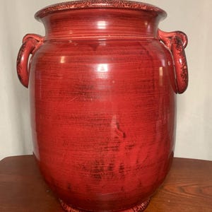 Puede incluir: Un jarrón grande de cerámica roja con dos asas en forma de lazo. El jarrón tiene un cuerpo ancho y redondeado y un borde ligeramente ensanchado. La pintura roja tiene un acabado envejecido, con matices negros visibles. Un artículo de decoración.