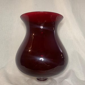 Puede incluir: Un jarrón de cristal rojo oscuro con un cuerpo bulboso y un borde acampanado. El jarrón se apoya sobre una base clara y circular. El cristal tiene un acabado brillante, que refleja la luz.