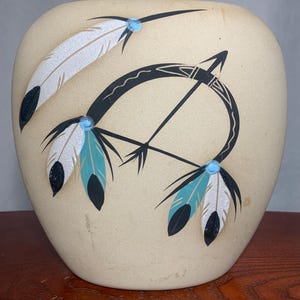 Op de afbeelding: Een beige keramische vaas met een afgeronde vorm. De vaas is versierd met een zwart pijl-en-boogontwerp, samen met witte en turquoise veren. De veren hebben zwarte accenten en kleine blauwe stippen.