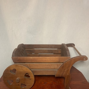 May include: A wooden toy wagon with a rectangular body and a curved handle. The wagon has a wheel with heart-shaped cutouts. The wood has a natural finish.